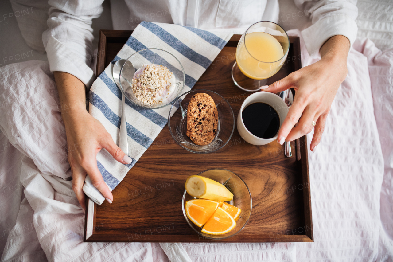 A midsection of woman lying in bed with coffee and breakfast indoors in the morning in a bedroom. A top view.