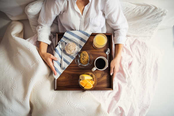 A midsection of woman lying in bed with coffee and breakfast indoors in the morning in a bedroom, using smartphone. A top view.