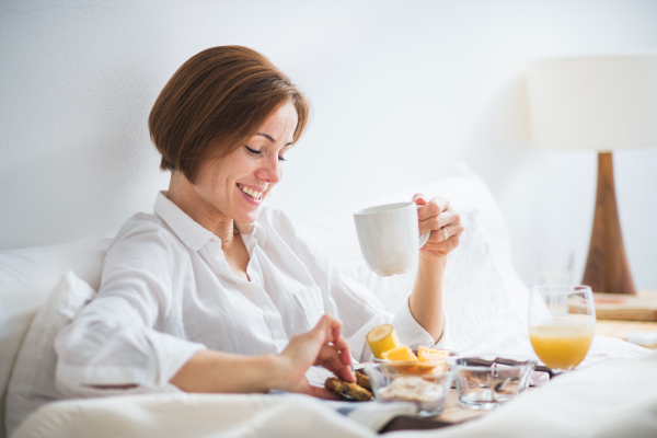 A young woman lying in bed with coffee and breakfast indoors in the morning in a bedroom.