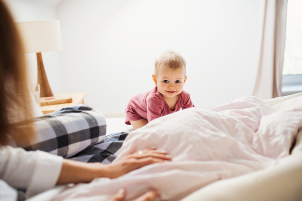 An unrecognizable young mother with little daughter sitting indoors on bed in the morning, playing.