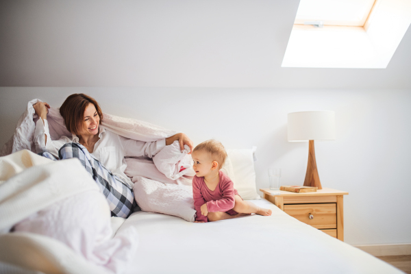 A happy young mother with little daughter sitting indoors on bed in the morning, playing.