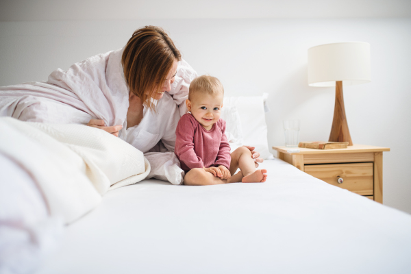 A happy young mother with little daughter sitting indoors on bed in the morning, playing.