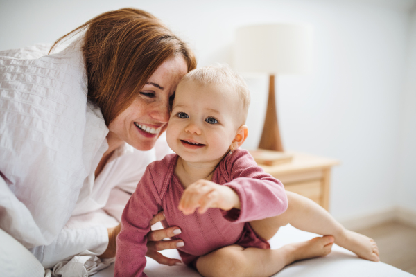 A happy young mother with little daughter sitting indoors on bed in the morning, playing.