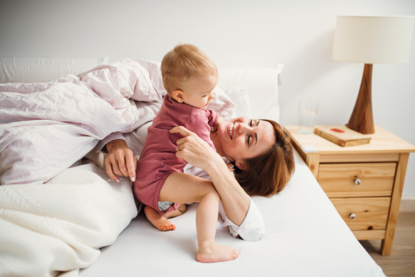 A happy young mother with little daughter sitting indoors on bed in the morning, playing.