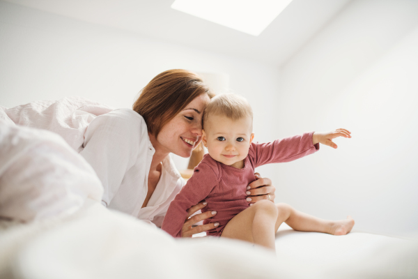 A happy young mother with little daughter sitting indoors on bed in the morning, playing.