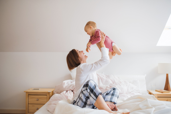 A happy young mother with little daughter sitting indoors on bed in the morning, playing.