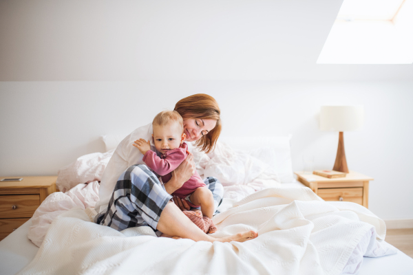 A happy young mother with little daughter sitting indoors on bed in the morning, playing.