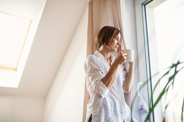 A young woman with night shirt standing by the window in the morning, looking out and holding a cup of coffee. Copy space.