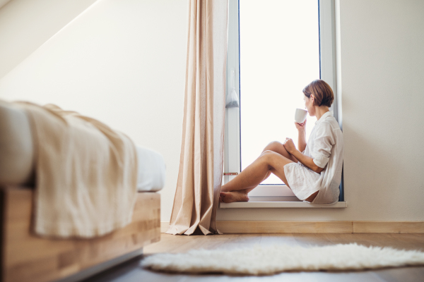 A young woman with night shirt sitting by the window in the morning, drinking coffee.