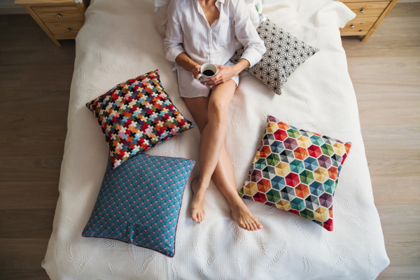 A midsection view of young woman with night shirt sitting indoors on bed in the morning, holding coffee.