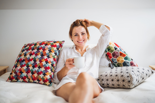 Young woman with night shirt sitting indoors on bed in the morning, holding cup of coffee.