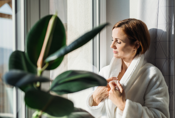 A young woman with bath robe and closed eyes standing indoors by a window in the morning, meditating. Copy space.