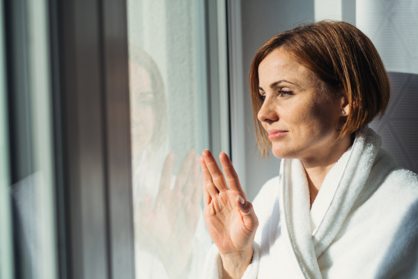 A young woman with bathrobe standing by the window in the morning, looking out. Copy space.