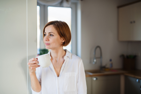 A young content woman with cup of coffee standing indoors, leaning on a wall. Copy space.