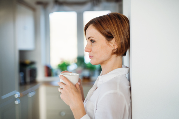 A young content woman with cup of coffee standing indoors, leaning on a wall. Copy space.