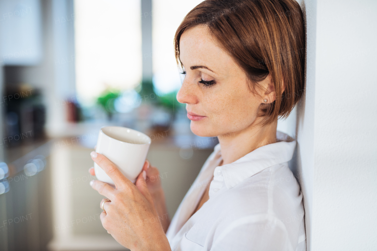 A young content woman with cup of coffee standing indoors, leaning on a wall. Copy space.