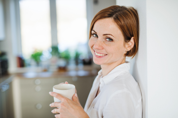 A young content woman with cup of coffee standing indoors, leaning on a wall. Copy space.