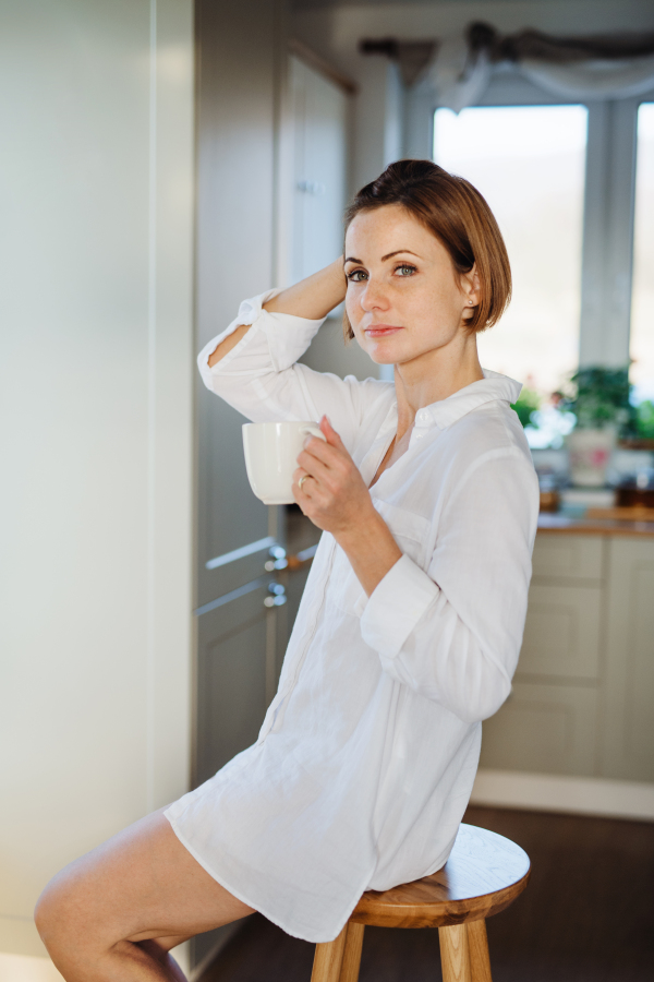 A young woman with cup of coffee sitting on a stool indoors, holding cup of coffee. Copy space.