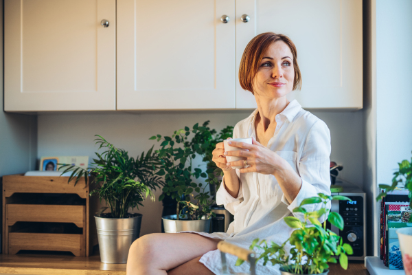 A young woman with white night shirt sitting on worktop indoors in kitchen, holding a cup of coffee.