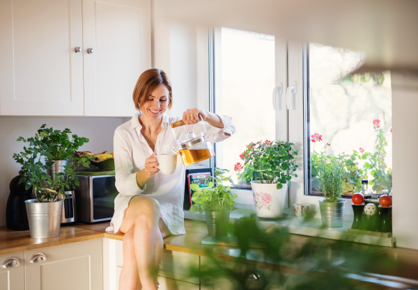 A young woman with white night shirt sitting on worktop indoors in kitchen, pouring tea.