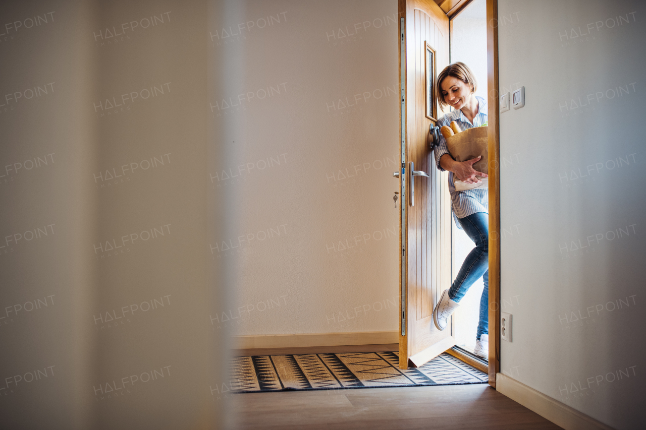 A young woman with groceries in paper shopping bag walking in through front door at home. Copy space.