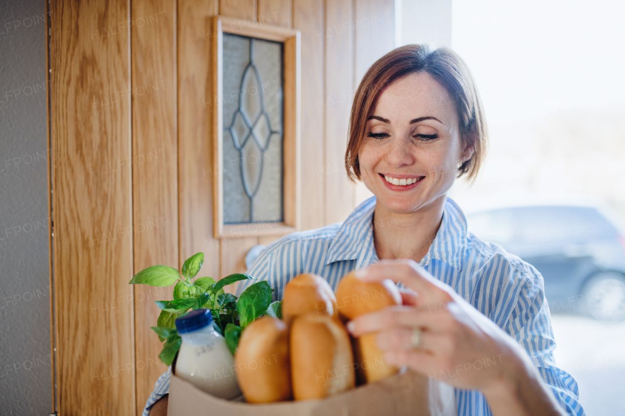 A young woman with groceries in paper shopping bag walking in through front door at home.