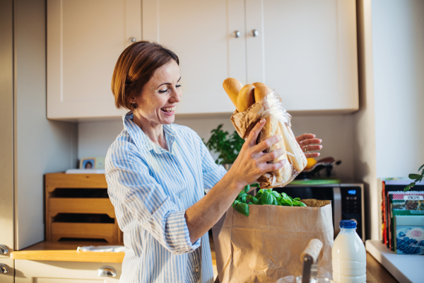 A young woman standing indoors in kitchen, unpacking groceries from shopping bag.