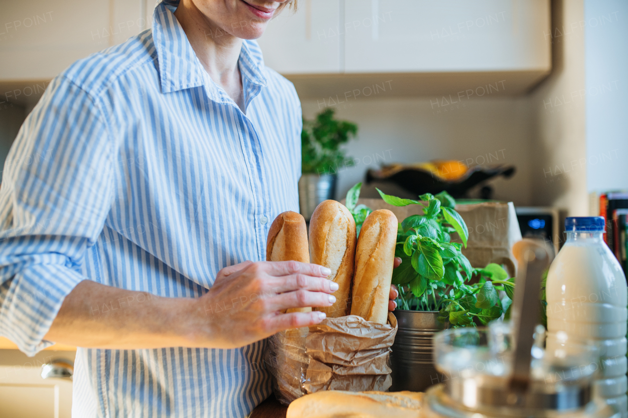 A midsection of young woman standing indoors in kitchen, unpacking groceries from shopping bag.