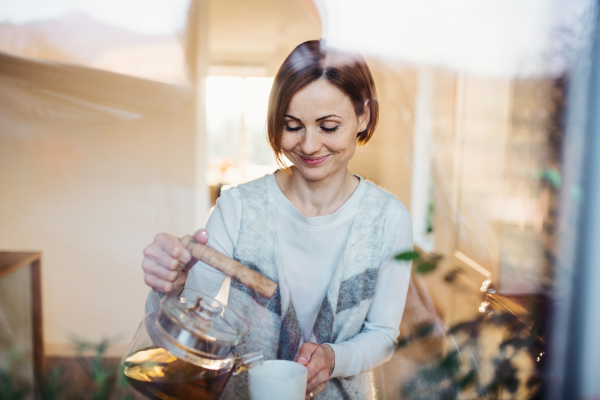 A happy young woman standing indoors in kitchen, pouring tea. Shot through glass.