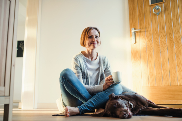 A young woman with a cup of coffee sitting indoors by the door on the floor at home, playing with a dog. Copy space.