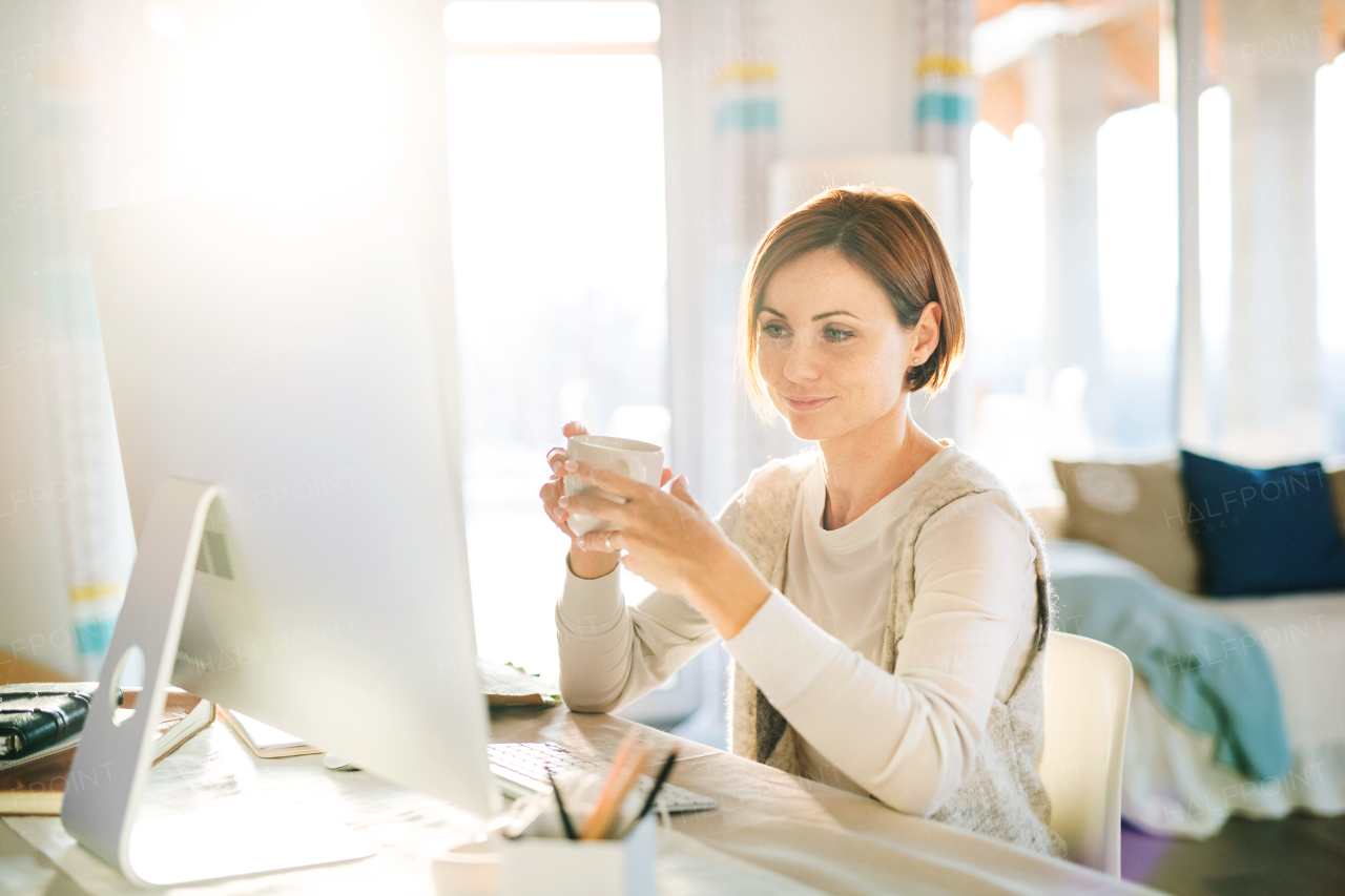 A happy young woman with smartphone, computer and coffee sitting at the desk indoors, working in a home office.