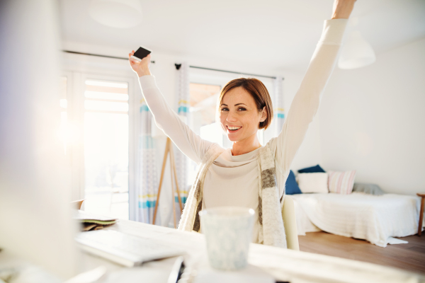 A happy young woman with outstretchced arms sitting indoors, working in a home office.