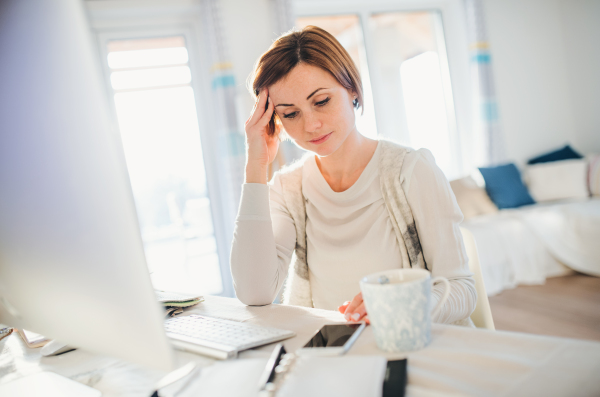 A tired young woman with smartphone sitting indoors, working in a home office.