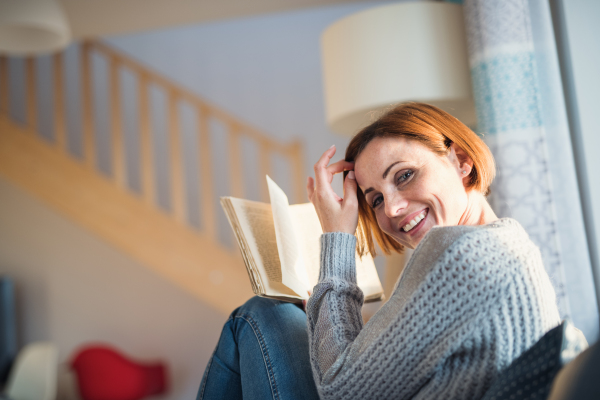 A cheerful young woman sitting indoors on a sofa at home, reading a book.