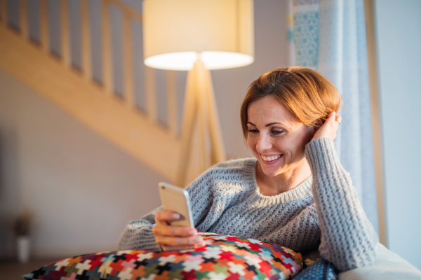 A happy young woman sitting indoors on a sofa in the evening at home, using smartphone.