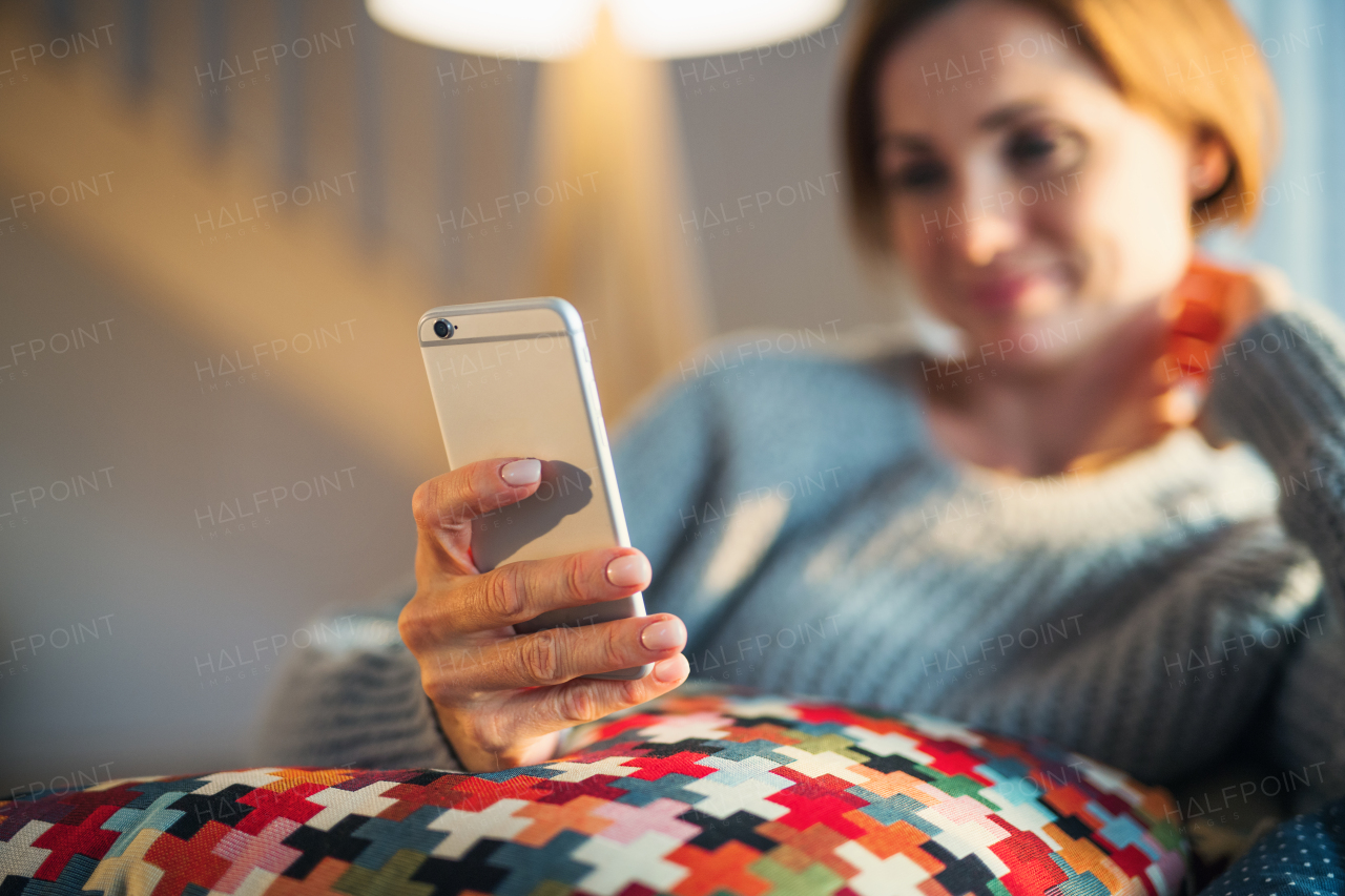 A happy young woman sitting indoors on a sofa in the evening at home, using smartphone.