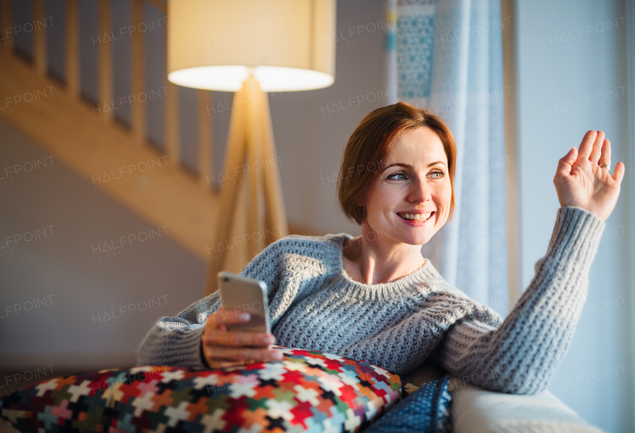 A happy young woman with smartphone sitting indoors on a sofa in the evening at home, waving at somebody.