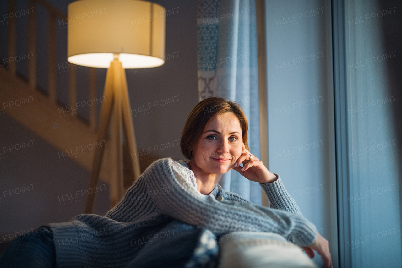 A happy young woman sitting indoors on a sofa in the evening at home, looking at camera.