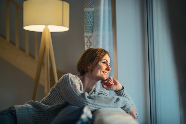 A happy young woman sitting indoors on a sofa in the evening at home, looking out of a window.