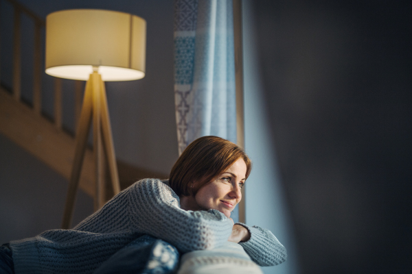 A happy young woman sitting indoors on a sofa in the evening at home, looking out of a window.