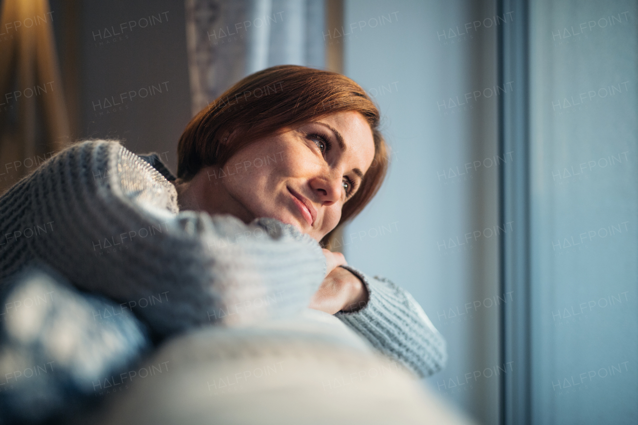 A happy young woman sitting indoors on a sofa in the evening at home, looking out of a window.