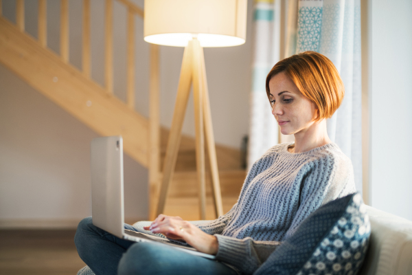 A happy young woman sitting indoors on a sofa in the evening at home, using laptop.