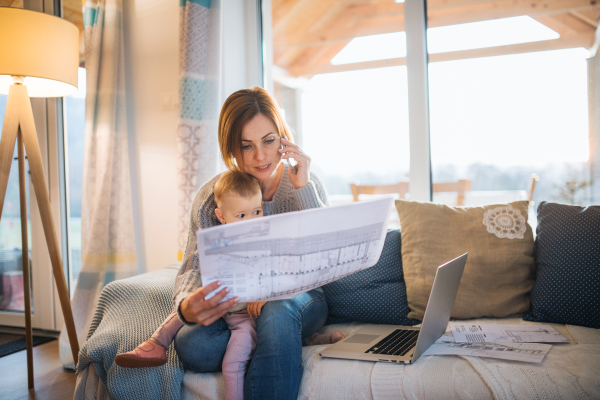 A young woman on the phone with a laptop, blueprints and toddler daughter sitting indoors, working in home office.