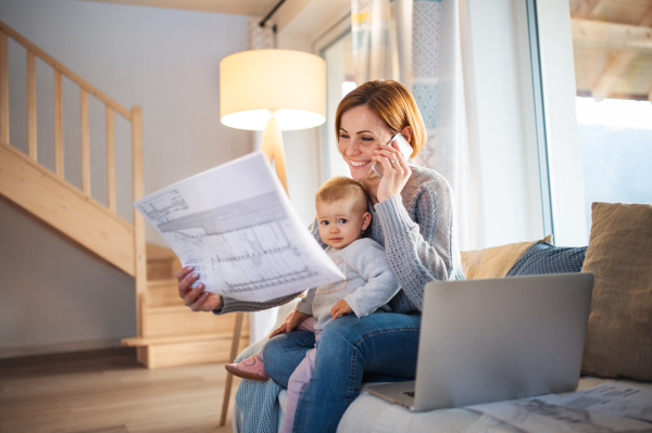 A young woman on the phone with a toddler daughter sitting indoors, working at home and holding blueprints.