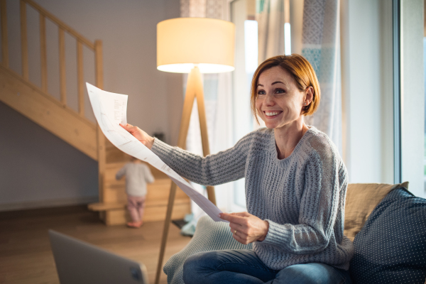 A young woman with laptop and blueprints sitting indoors, working at home.