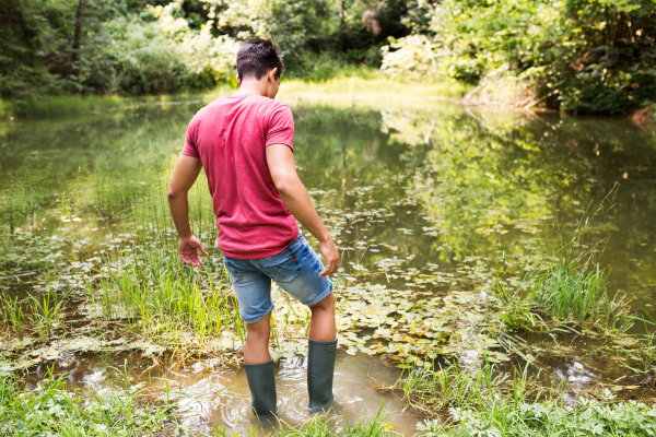 Teenage boy in rubber boots standing in lake. Summer vacation adventure. Rear view.