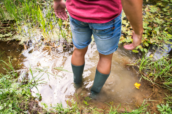 Unrecognizable young man in rubber boots standing in the lake. Summer vacation adventure. Rear view.