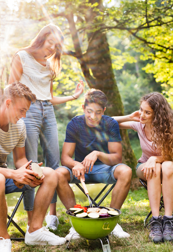 Beautiful teenagers enjoying camping vacations in forest, cooking vegetables on barbecue grill.