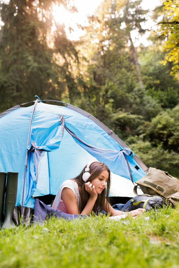 Two teenage girls camping in forest, sitting in front of tent, reading a book and listening to music. Summer vacation adventure.