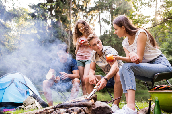 Beautiful teenagers enjoying camping vacations in forest. Boys cooking meat on bonfire.